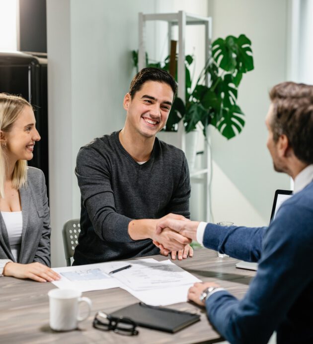 Happy couple shaking hands with their real estate agent at home. Young happy couple making an agreement with their financial advisor at home. Men are shaking hands.